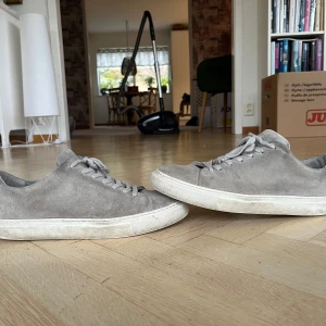 Grå sneakers från Sneaky Steve - Snygga grå sneakers i mocka från Sneaky Steve. Skorna har en klassisk design med snörning och en vit sula. Perfekta för en stilren look.
