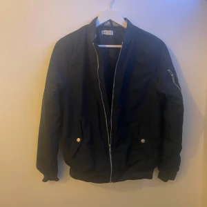 Svart bomberjacka med dragkedja - Snygg svart bomberjacka med dragkedja framtill och fickor med knappar. Jackan har en klassisk design med ribbade muddar och en ficka med dragkedja på ärmen. Perfekt för en stilren look. Pris är förhandlingsbart 