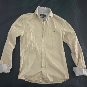 Beige skjorta från Morris - Snygg skjorta från Morris med logga broderad på bröstet. Skjortan är i bra skick och passar perfekt in till våren och sommarn. Skjortan sitter i storlek S och modellen är ca 178 cm lång. Hör gärna av er vid frågor eller funderingar!
