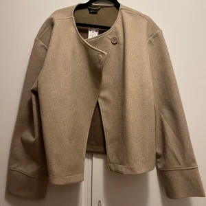 Beige kavaj med en knapp - Säljer en stilren beige kappa med en knapp framtill. Den säljer jag pga att jag köpte den i en annan storlek, man kan använda jackan på två olika sätt
