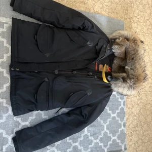 Svart parkajacka från Parajumpers - Säljer en svart parkajacka från Parajumpers med pälsfodrad huva och flera praktiska fickor. Jackan har både dragkedja och knappar framtill samt en snygg detalj med gul rem vid kragen. Perfekt för kalla dagar!