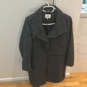 Grå kappa från Isolde - Snygg grå kappa från Isolde med stilrena detaljer. Den har en hög krage och knappar framtill. Perfekt för kyligare dagar och ger en elegant look. Två praktiska fickor med dragkedja på framsidan.