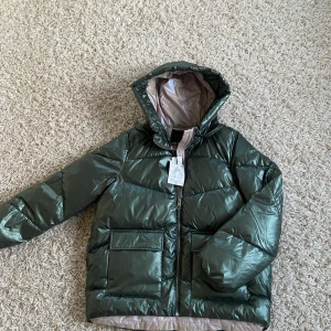 Grön pufferjacka från Mohito - Snygg grön pufferjacka från Mohito med glansig finish och dragkedja framtill. Jackan har en praktisk huva och stora fickor, perfekt för kyliga dagar. Insidan är ljus och kontrasterar snyggt mot den gröna utsidan.
