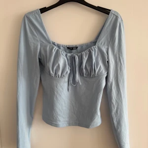 Ljusblå långärmad topp med knyt - Säljer en ljusblå långärmad topp med knytning framtill. Toppen har en åtsittande passform och en söt detalj vid bysten. Perfekt för en stilren look.