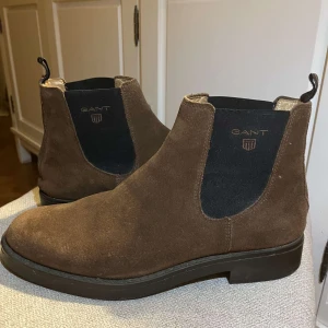 Bruna mocka Chelsea boots från Gant - Snygga bruna Chelsea boots från Gant i mjuk mocka. De har en klassisk design med elastiska paneler på sidorna för enkel på- och avtagning. Perfekta för en stilren look. Säljer dem för att jag inte använder dem längre. Kostar ca 2700 kr nya. Skick 9/10 knappt använda! Hör av er ifall ni har några funderingar mvh Albin! Ps! Stora i storleken