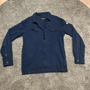 Mörkblå skjorta från Cars Jeans - Snygg mörkblå skjorta från Cars Jeans med dragkedja framtill och två bröstfickor. Skjortan har långa ärmar och en cool patchdetalj på ena ärmen. Perfekt för en avslappnad stil.