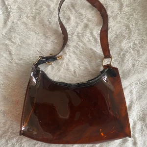 Transparent brun axelväska - Snygg transparent brun axelväska med dragkedja. Väskan har en modern design med ett brett axelband och är perfekt för att bära med sig det nödvändigaste. Den genomskinliga looken ger en unik touch.