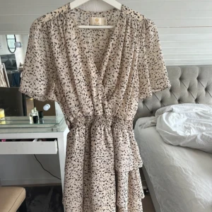 Prickig omlottklänning - Säljer en chic beige omlottklänning med svart prickigt mönster. Klänningen har korta ärmar och en smickrande v-ringad design. Klänningen är i strl M/38🙌🏼