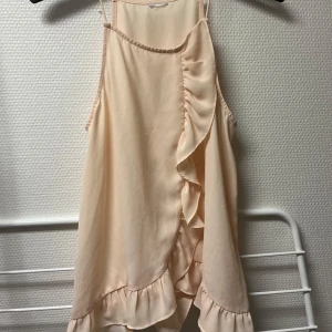 Beige/rosa volanglinne från Gina Tricot - Snyggt beige linne från Gina Tricot med volangdetaljer framtill och vid fållen. Tillverkat i 100% polyester, perfekt för sommarens varma dagar. 