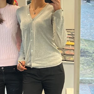 Grå kofta från Filippa K - Säljer en stilren grå kofta från Filippa K i 100% bomull. Koftan har en v-ringad design med knappar framtill och långa ärmar. Har ett hål i armhålan men märks knappt när man har den på. Skulle säga att den är mer S än M💞