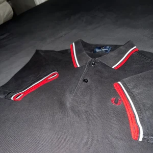 Fredperry piké - Snygg svart pikétröja från Fred Perry med klassiska röda och vita ränder på krage och ärmslut. Storlek XS men passar även S . Tillverkad i 100% bomull, ett hål vid sömmen på ena ärmen. Ny pris ca 500kr