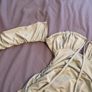 Beige offshoulder klänning - Trendig beige klänning med offshoulder-design och långa ärmar. Klänningen har en smickrande passform med knytband och volangdetaljer vid axlarna. Perfekt för en stilfull look.