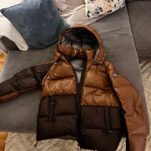 Brun och svart pufferjacka från Colmar - Säljer en snygg brun och svart pufferjacka från Colmar. Jackan har en hög krage och en praktisk dragkedja framtill. Den är perfekt för kyliga dagar med sin quiltade design och långa ärmar. En stilren och varm jacka för vintern! Ny pris på denna är 8999kr och den e knappt använd 