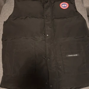 Svart dunväst från Canada Goose - Säljer en svart dunväst från Canada Goose då jag använt bara 1 gång,  med knappar och dragkedja. Västen har en klassisk design med två stora fickor framtill och en broderad logotyp på bröstet. Perfekt för våren och hösten.