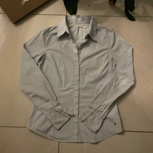 Ljusblå skjorta från Tommy Hilfiger - EliteVolt.  Snygg ljusblå skjorta från Tommy Hilfiger med klassisk krage och knappar framtill. Skjortan har långa ärmar och ett diskret broderat märke vid nederkanten. Perfekt för en stilren look.