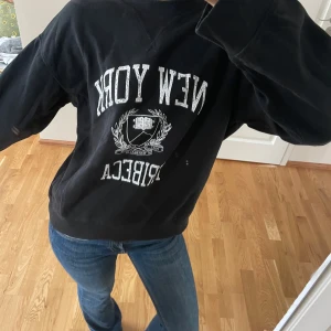 Svart sweatshirt med New York-tryck - Säljer en svart sweatshirt med texten 'New York Tribeca' i vitt. Tröjan har långa ärmar och en avslappnad passform, perfekt för en casual look. Den har ett tryck med ett emblem i mitten. Passar bra till jeans eller andra avslappnade outfits.