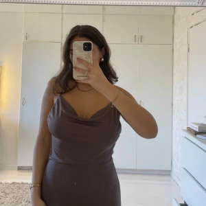 Brun klänning med draperad hals - Snygg brun klänning med draperad hals och smala axelband. Perfekt för en elegant look. Klänningen har en figurnära passform och når till vaderna. 