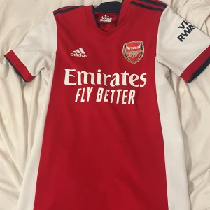 Röd Arsenal fotbollströja från Adidas - Säljer en röd Arsenal fotbollströja från Adidas med vita ärmar och klubbens emblem på bröstet. 
