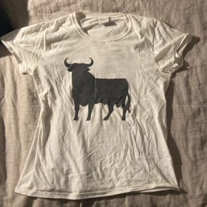 Vit t-shirt med tjurmotiv från Toro - Säljer en vit t-shirt från Toro med ett stort svart tjurmotiv på framsidan. T-shirten är kortärmad och tillverkad i bomull, perfekt för en avslappnad stil. Har två små hål vid ena armhålan.