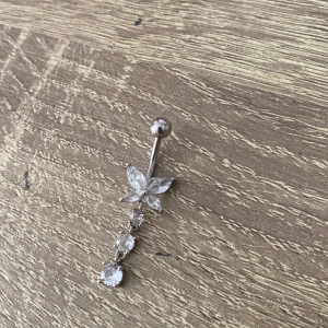 Silverfärgat navelpiercing med en fjäril och 3 st cirklar under, använd 4 gånger.  - Elegant navelpiercing i silverfärg med ett vackert blommotiv och hängande kristaller. Perfekt för att ge en glittrig touch till din stil.