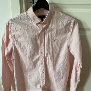 Ljusrosa skjorta från GANT -  Ljusrosa skjorta från GANT. Skjortan har långa ärmar och knappar framtill samt en broderad logga på bröstet. .