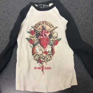Vit och svart långärmad tröja från Bershka - Säljer en cool vit och svart långärmad tröja från Bershka med Guns N' Roses-tryck. Tröjan har ett stort, färgglatt motiv med rosor och dödskallar på framsidan. Perfekt för rockälskare! 🎸