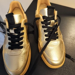 Guldiga sneakers med svarta detaljer - Säljer ett par snygga guldiga sneakers med svarta snören och detaljer. De har en cool, glänsande finish och en bekväm sula som ger bra grepp. Perfekta för att sticka ut i mängden! märket Tamaris kvitto finns nypris 1399 kr.