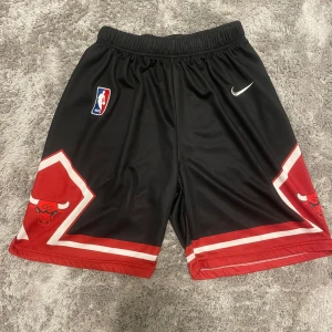 Svarta och röda basketshorts med NBA-logga - Säljer ett par snygga basketshorts i svart och rött från Nike. De har en elastisk midja och är dekorerade med en NBA-logga och ett tjurmotiv på sidorna. Perfekta för basketträning eller bara för att chilla i. 🏀