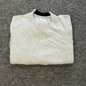 Grå sweatshirt från C.P. Company - Säljer en stilren grå sweatshirt från C.P. Company. Den har en cool detalj med en svart lins på ärmen och en dragkedja. Perfekt för en casual look och passar till det mesta. Tröjan är i bra skick och superbekväm!