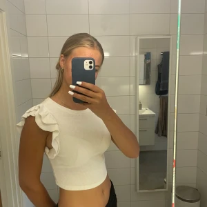 Vit ribbad topp med volangdetaljer - Säljer en superfin vit ribbad topp med volangdetaljer på axlarna. Den är croppad och har en tight passform, perfekt för sommaren. Toppen är ärmlös och gjord i ett mjukt bomullsmaterial. Passar både till vardags och fest!