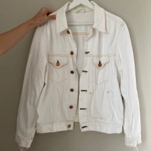 Vit jeansjacka från Acne - Säljer en snygg vit jeansjacka från Acne. Den har en klassisk design med brons knappar och kontrasterande sömmar i gult. Jackan har två bröstfickor med lock och knappdetaljer. Perfekt för vår och sommar! Säljer pga knappt använd.