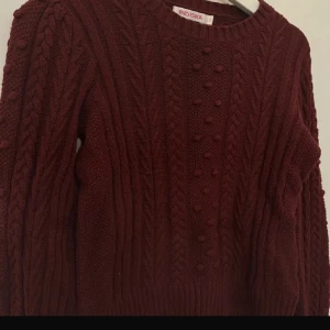 Vinröd stickad tröja från Indiska - Säljer en supermysig vinröd stickad tröja från Indiska. Den har ett snyggt flätmönster och små bollar på framsidan som ger en extra touch. Perfekt för kalla höst- och vinterdagar. Tröjan har långa ärmar och ribbstickade muddar vid ärmslut och nederkant. Passar perfekt till både jeans och kjol!