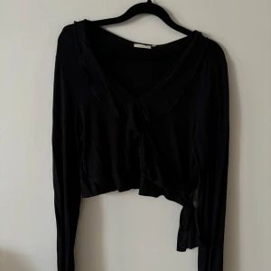 Svart långärmad kofta från Fransa - Säljer en svart topp från Fransa i storlek L, men liten i storleken. Den har långa ärmar och en knytning framtill som ger en fin detalj. Perfekt för höst och vinter, och kan enkelt matchas med olika outfits. 