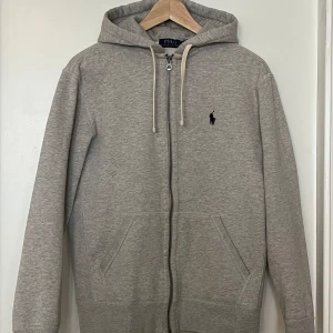 Grå hoodie från Polo Ralph Lauren - Säljer en snygg och bekväm grå hoodie från Polo Ralph Lauren. Den har en dragkedja framtill och en klassisk logga på bröstet. Perfekt för året om, med en skön passform. Den är i bra mycket bra skick.