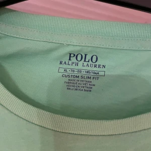 Grön t-shirt från Polo Ralph Lauren - Säljer en fräsch grön t-shirt från Polo Ralph Lauren i storlek XL. Den har en custom slim fit passform och är tillverkad i mjuk bomull. Perfekt för sommardagar eller avslappnade tillfällen. Den klassiska loggan är broderad i orange på bröstet.