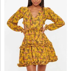 Missguided gul klänning öppen rygg - En gul klänning med blommönster och långa ärmar. Klänningen har en urringning framtill och en öppen rygg med snörning. Den har även en volangdetalj i nederkanten.