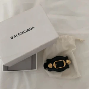 Svart armband från Balenciaga - Snyggt svart armband från Balenciaga med gulddetaljer. Armbandet har en unik design med ett spänne i mitten och är tillverkat i skinn. Perfekt för att ge en lyxig touch till vilken outfit som helst. Kommer i originalförpackning.