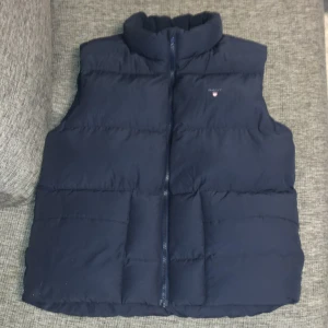 Mörkblå dunväst från GANT - Säljer en snygg mörkblå dunväst från GANT i storlek 170 cm, perfekt för kyliga dagar. Västen har en dragkedja framtill och är i nyskick. Den är stilren och passar bra till både vardag och fest. Ett måste för garderoben! 💙 Ny pris 1500kr💙