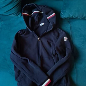 Mörkblå  huvtröja från Moncler - Säljer en stilren svart huvtröja från Moncler med dragkedja och broderad logga på ärmen. Tröjan har en huva med röd och vit randig detalj. Den är i nyskick knappast använd. Alla qr coder och sånt fungerar såklart.