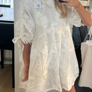 Vit blommig klänning - Snygg vit klänning med blommigt mönster och puffiga kortärmar. Klänningen har en lös passform och knytband vid ärmarna för extra detalj. Perfekt för sommaren eller studenten. Helt oanvänd med prislappen kvar!