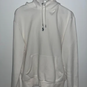 Vit hoodie från Ralph Lauren - Säljer en stilren vit hoodie från Ralph Lauren med klassisk känguruficka och justerbar huva. Den har en liten broderad logga på bröstet och är perfekt för en avslappnad look. Skick 9/10