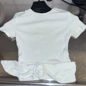Vit ribbad topp från Bershka - Säljer en vit ribbad topp från Bershka i storlek XS. Toppen har korta ärmar och en knytning i midjan som ger en snygg detalj. Perfekt för en stilren look.