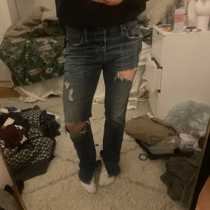 Blå jeans med slitningar - Säljer ett par blå jeans med coola slitningar på knäna och låren. De har en klassisk femficksdesign och en bekväm passform. Perfekta för en avslappnad stil.