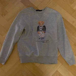 Grå sweatshirt med Polo Bear från Ralph Lauren - Säljer en grå sweatshirt från Ralph Lauren med det ikoniska Polo Bear-motivet på framsidan. Tröjan har långa ärmar och en rund halsringning. Perfekt för en avslappnad stil. Obs.  Den är lite sliten på björnen