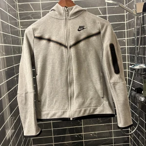 Grå hoodie från Nike - Säljer en stilren grå hoodie från Nike med dragkedja och svart logga på bröstet. Jackan har långa ärmar och en praktisk ficka på ärmen. Perfekt för en sportig look eller avslappnade dagar.den passar nog mellan 160 168 