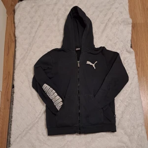 Svart huvtröja från Puma - Säljer en svart huvtröja från Puma med dragkedja och logga på bröstet. Tröjan har långa ärmar och en stor ficka framtill. Perfekt för en avslappnad stil.