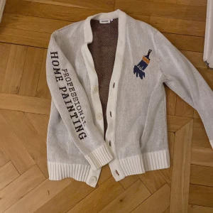 Vit kofta från Weekday - Säljer en vit kofta från Weekday med texten 'Professional Home Painting' och en broderad pensel på framsidan. Koftan har knappar och ribbade muddar. Perfekt för en unik stil.