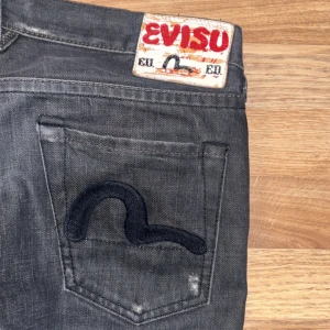 Svarta jeans från Evisu - Snygga svarta jeans från Evisu med det ikoniska logotypen broderad på bakfickorna. Jeansen har en klassisk femficksdesign och en knappgylf. Professionellt sydda kortare då de var för långa på mig. Svart/grå distessed färg.