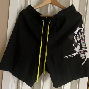 Svarta shorts med grafiskt tryck - Snygga svarta shorts från Firefly med coolt grafiskt tryck och dödskallemotiv. De har en elastisk midja med gula snören för justering. Perfekta för en avslappnad stil.