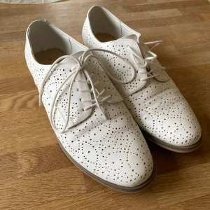 Vita perforerade skor med snörning - Snygga vita skor med perforerat mönster och snörning. De har en låg klack och en spetsig tå, vilket ger en elegant touch. Perfekta för en stilren look.